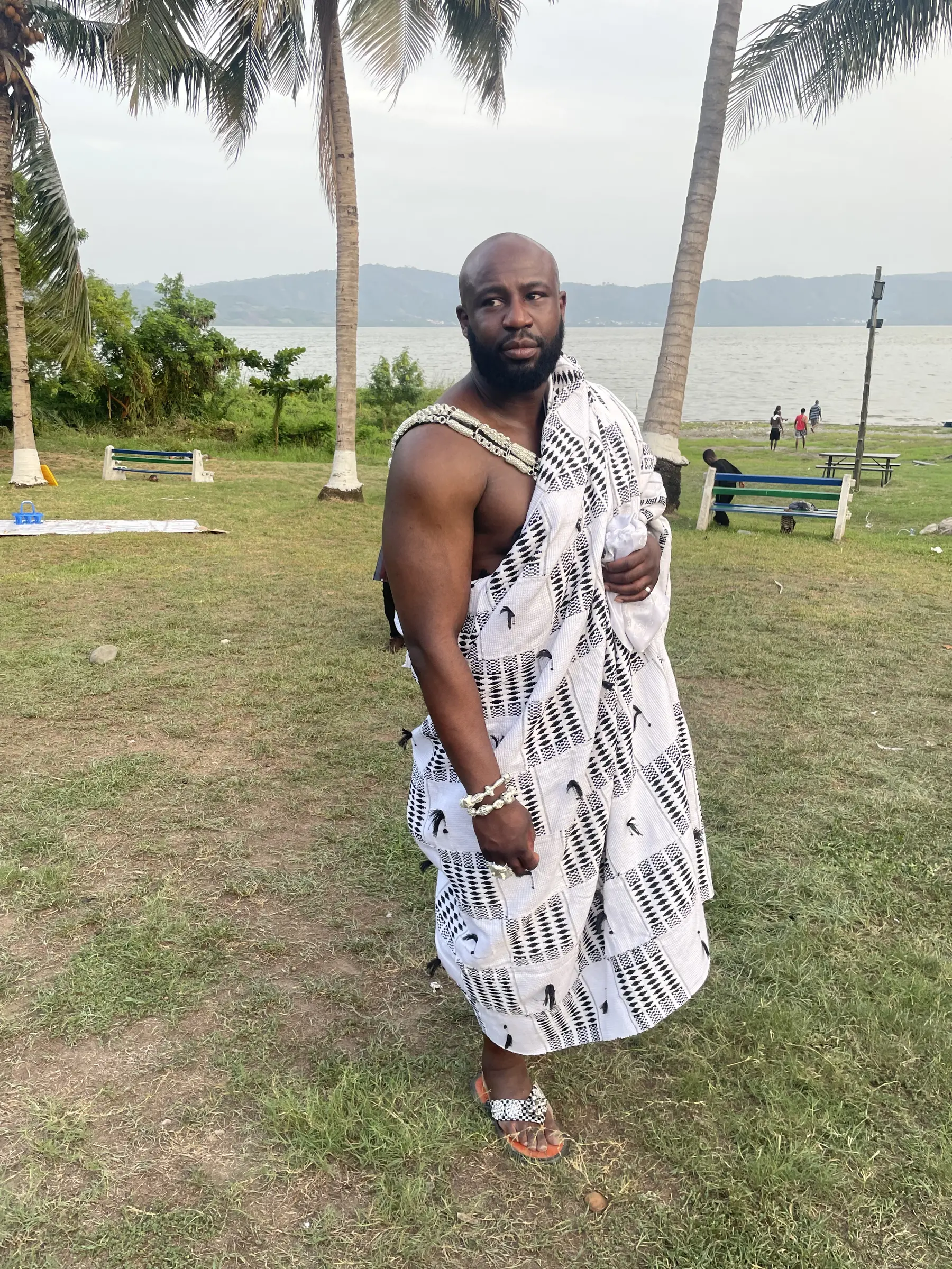 Dennis Toppin in adinkra-doek met kralen en chief-sandalen, staand aan de oever van Lake Bosomtwe onder palmen.