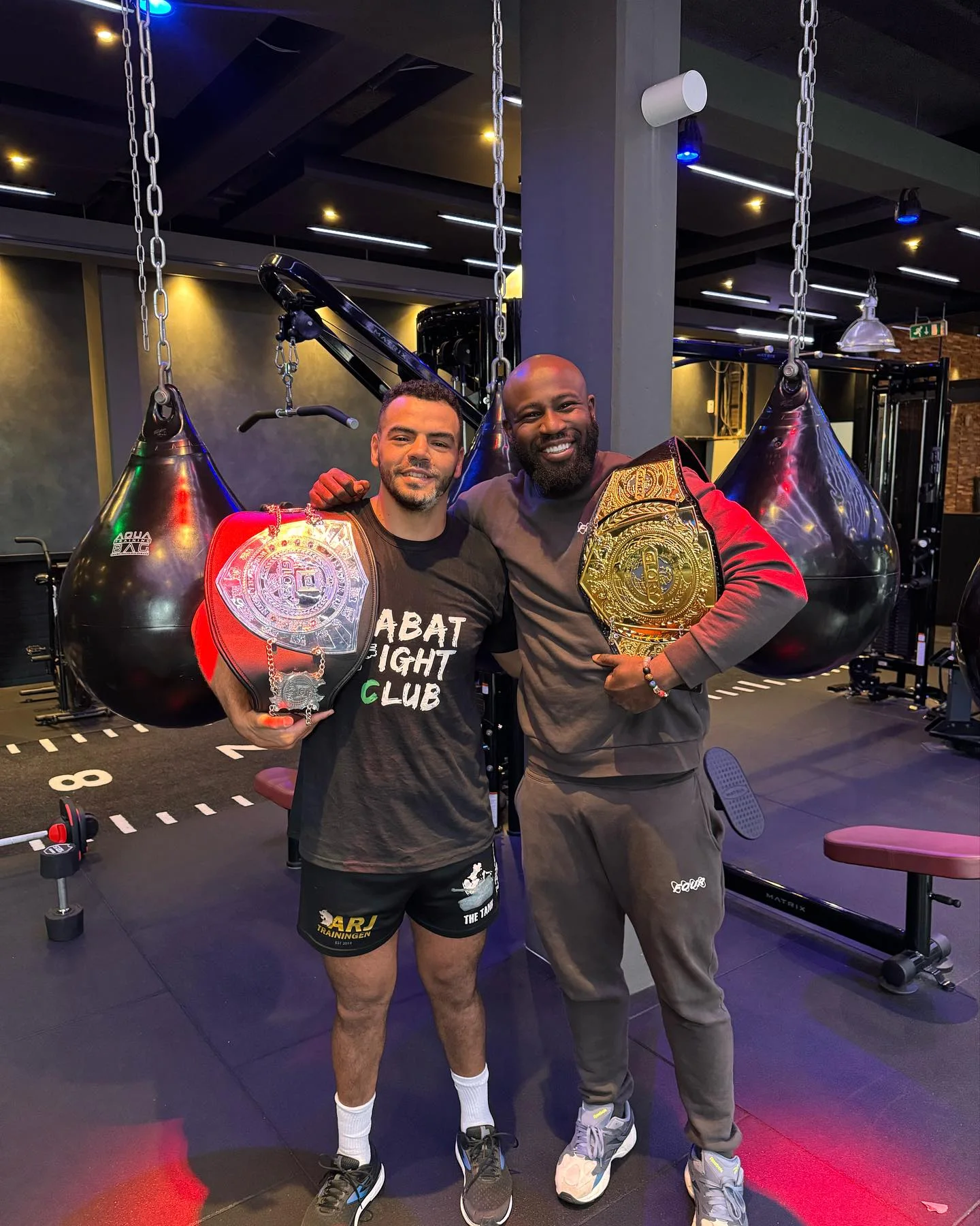 Dennis Toppin in de sportschool met een vechtsporter die twee kampioensriemen vasthoudt, staand tussen de bokszakken.