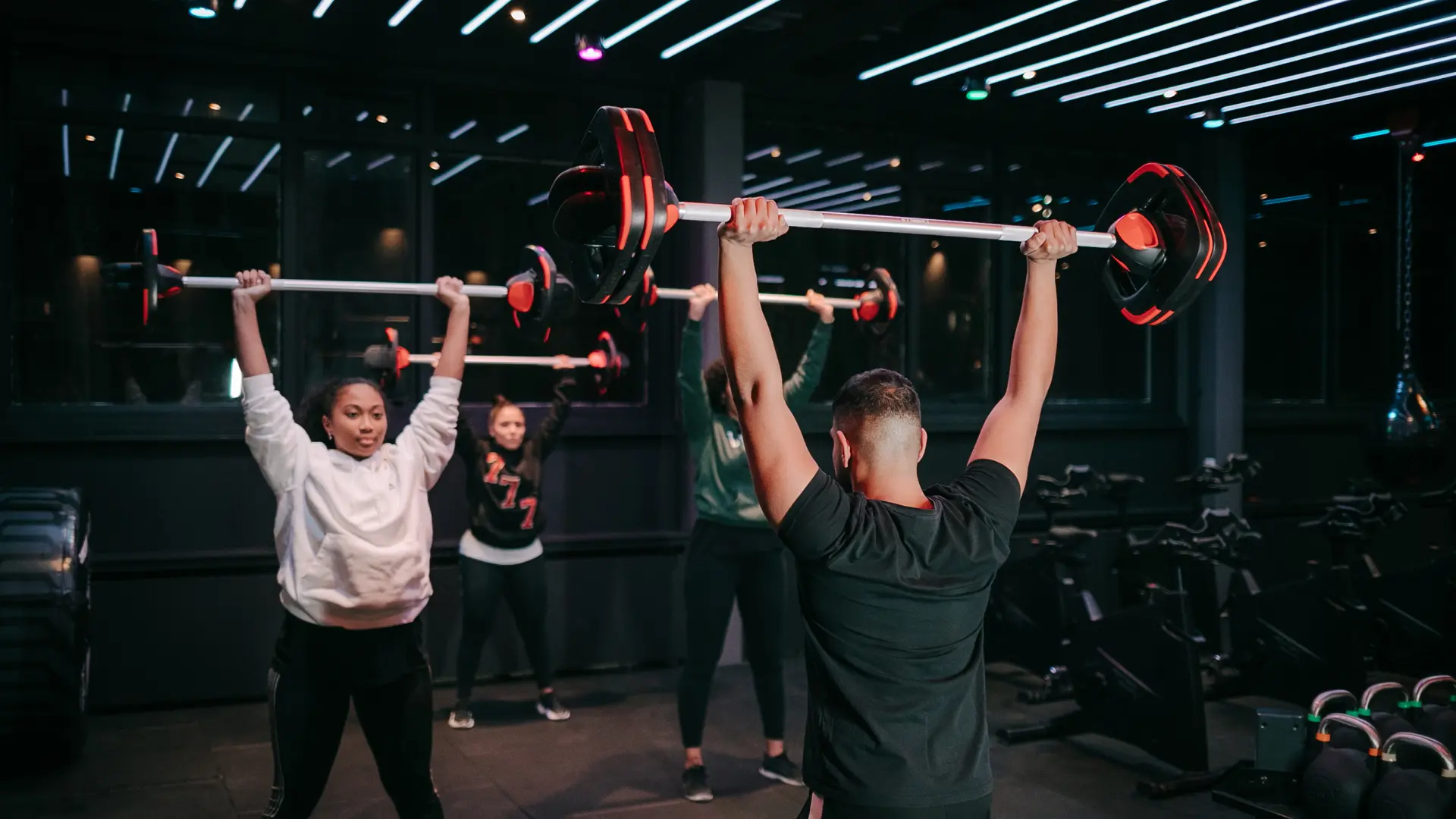 Een groepsles in de studio: meerdere mensen drukken tegelijk een halter boven het hoofd, met rode verlichting op de achtergrond.